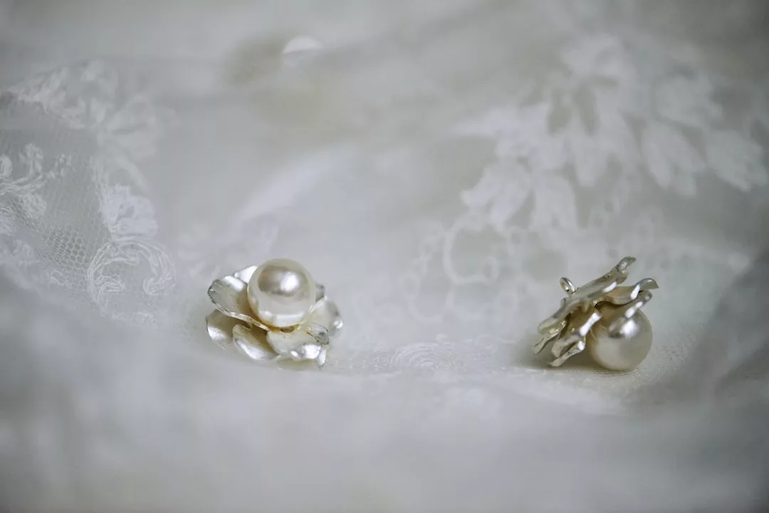 Two silver earrings with pearl-like centers rest on delicate white lace fabric, adding a touch of Glamesia fashion to your lifestyle.