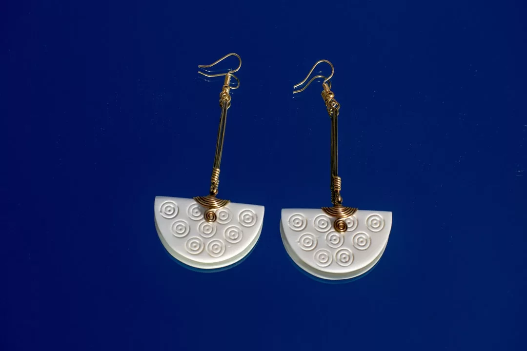 Pair of gold wire hook earrings with white semicircular pendants, decorated with embossed circular patterns, displayed on a dark blue background—a true statement of luxury and Glamesia fashion.