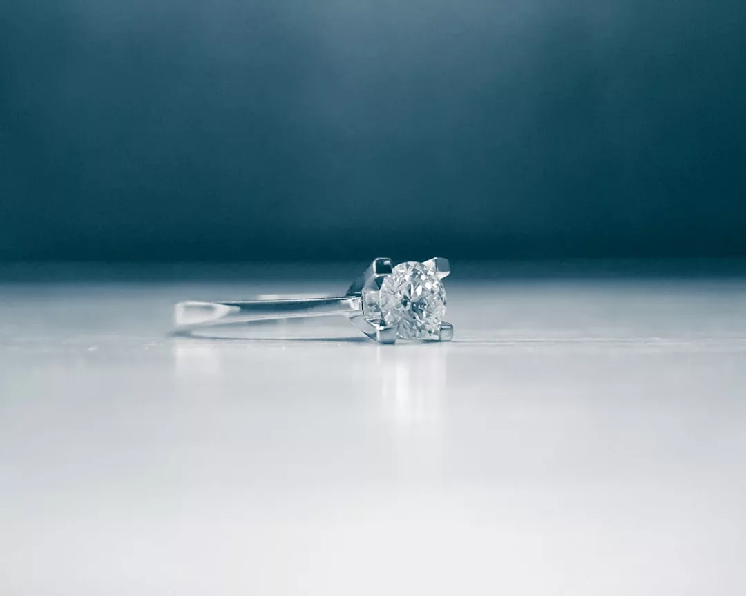 A close-up of a Glamesia silver ring with a single round diamond set in a prong setting, placed on a smooth, reflective surface with a blurred blue-gray background, capturing the essence of luxury.