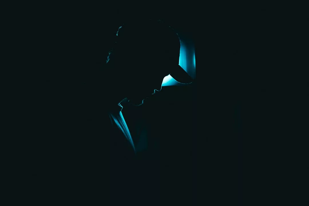 Silhouette of a person wearing a cap, illuminated from behind by a blue light in a dark environment, capturing an enigmatic fashion lifestyle.