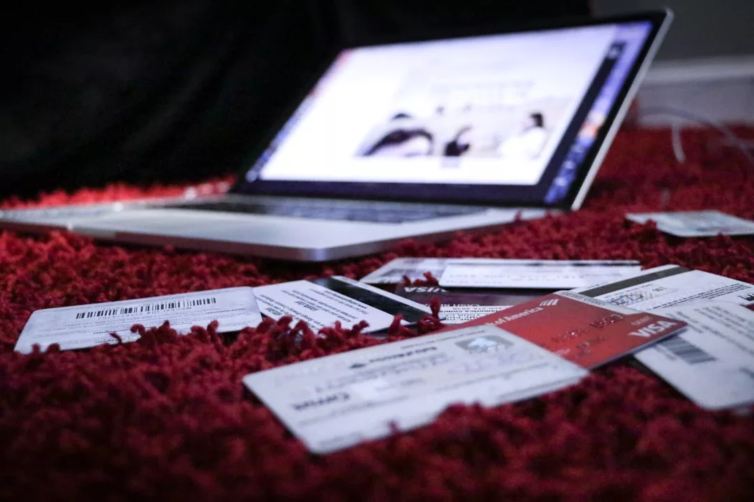 Several credit cards and other cards are scattered on a red carpet in front of an open laptop computer, reflecting a glimpse of Glamesia’s chic lifestyle.