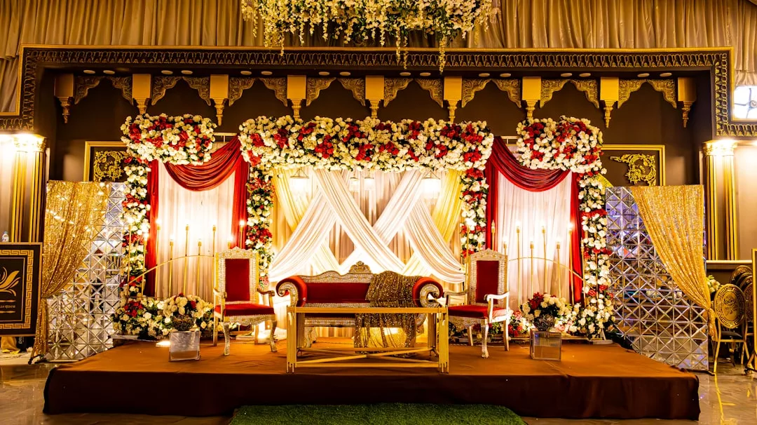 A decorated wedding stage with a red and gold sofa, red chairs, floral arrangements, draped fabric, and gold accents exudes Glamesia fashion in an ornate indoor venue.