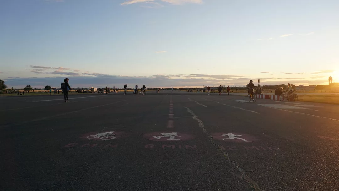 People walk, cycle, and skate on a wide, open paved path at sunset, with distance markings and symbols painted on the ground in the foreground—a scene where urban life meets effortless fashion.