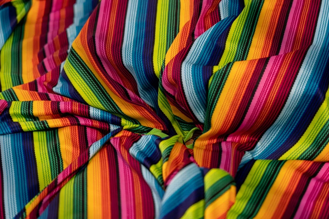 Colorful striped fabric from Glamesia featuring bright, parallel lines in red, pink, yellow, blue, green, and orange, gathered and creased in the center.