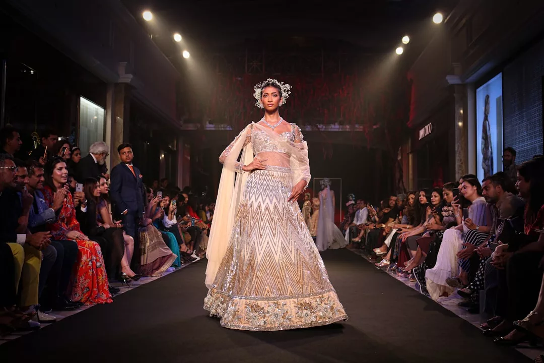 A model walks down the runway at a Glamesia fashion show, wearing an ornate, traditional outfit with a long skirt, sheer dupatta, and floral headpiece, as the audience watches in awe.