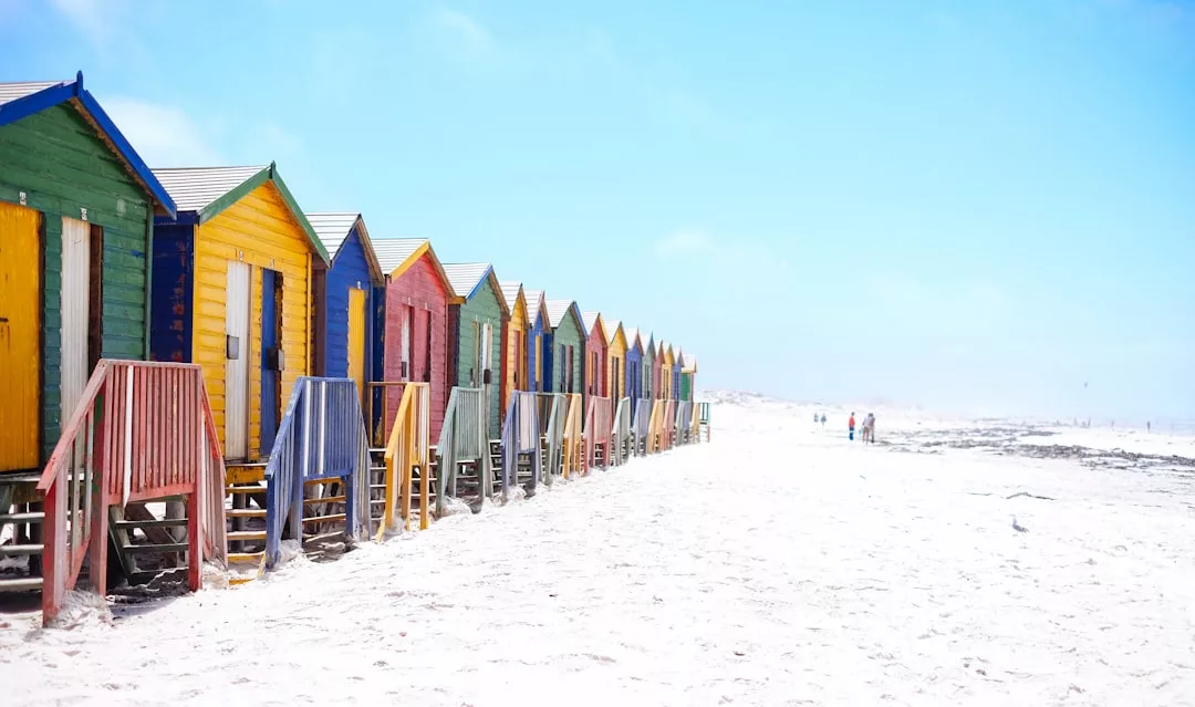 A row of colorful beach huts stands on a white sandy beach under a bright blue sky, reflecting a vibrant lifestyle, with a few people visible in the distance.