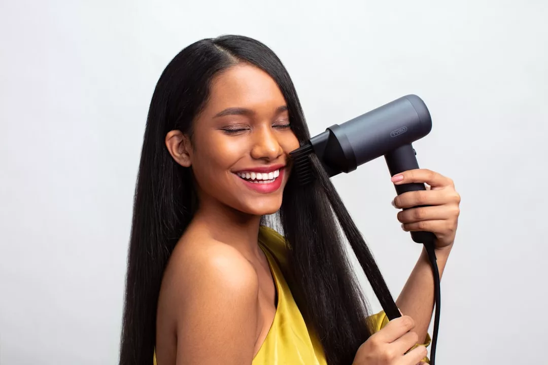 A woman with long, straight black hair uses a luxury black hair dryer and brush to style her hair, smiling with her eyes closed.