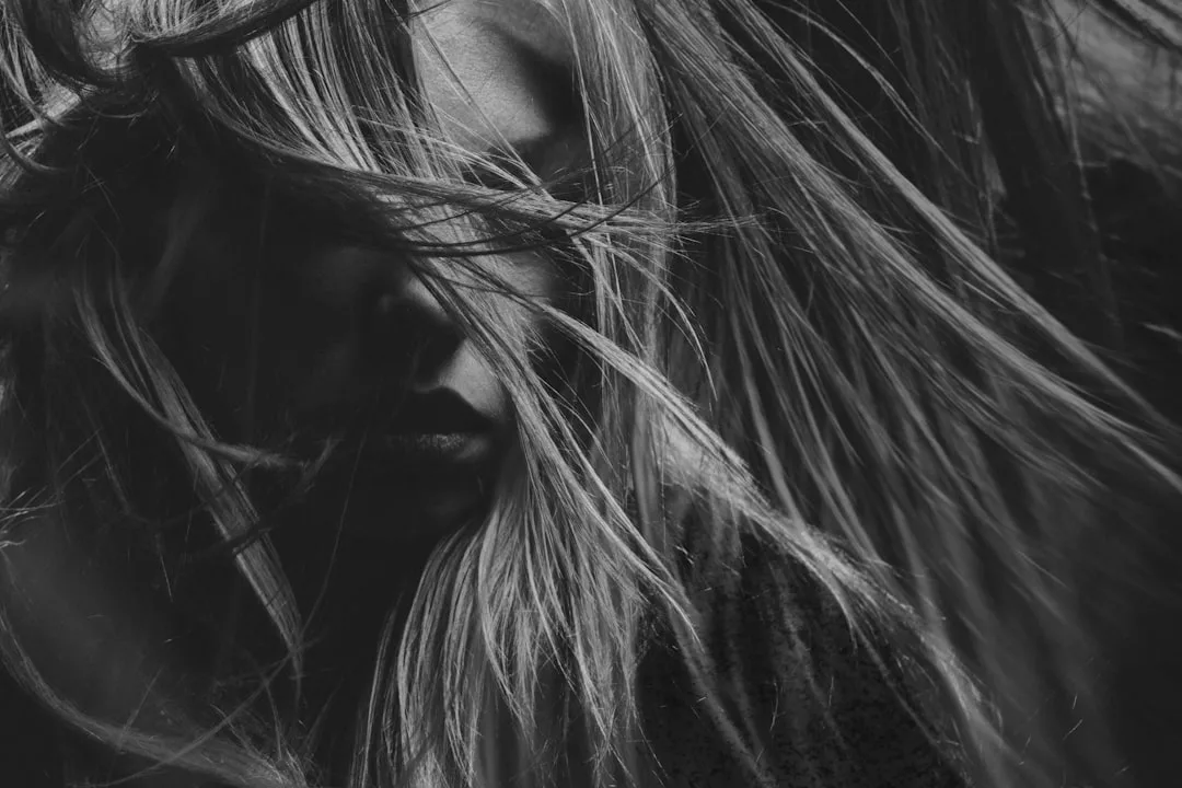 Black and white close-up of a person with long hair obscuring most of their face, capturing a dramatic, shadowed effect that hints at an enigmatic lifestyle.