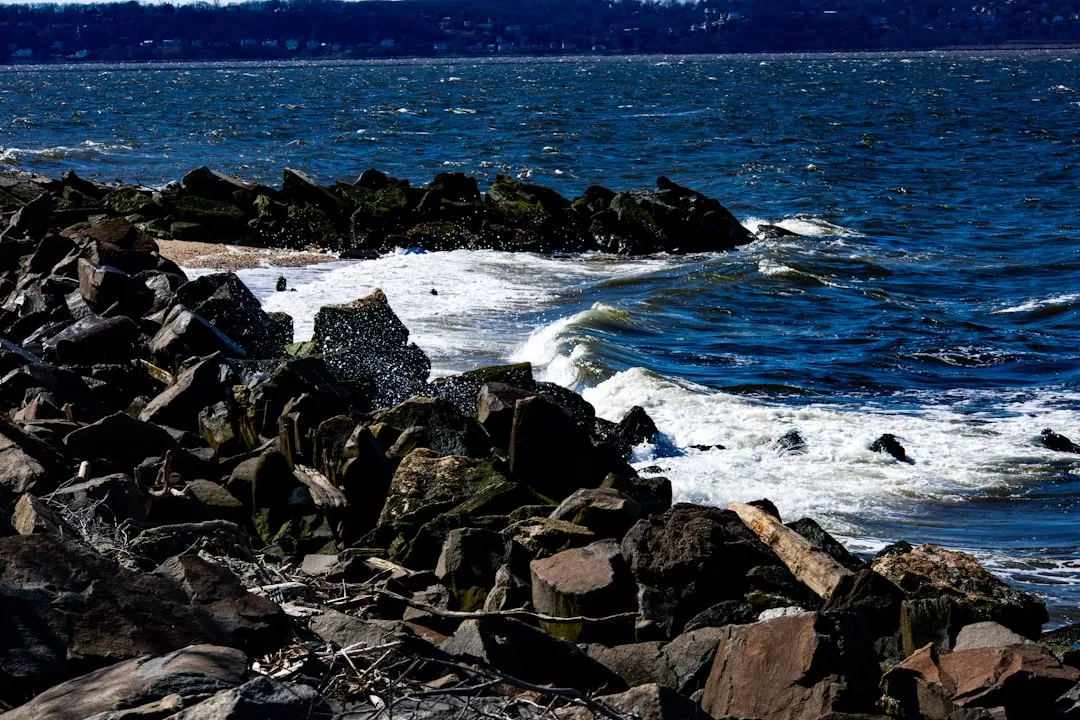 Waves crash against large rocks along a shoreline under a cloudy sky, with distant land visible across the water, capturing the essence of Laxary lifestyle.