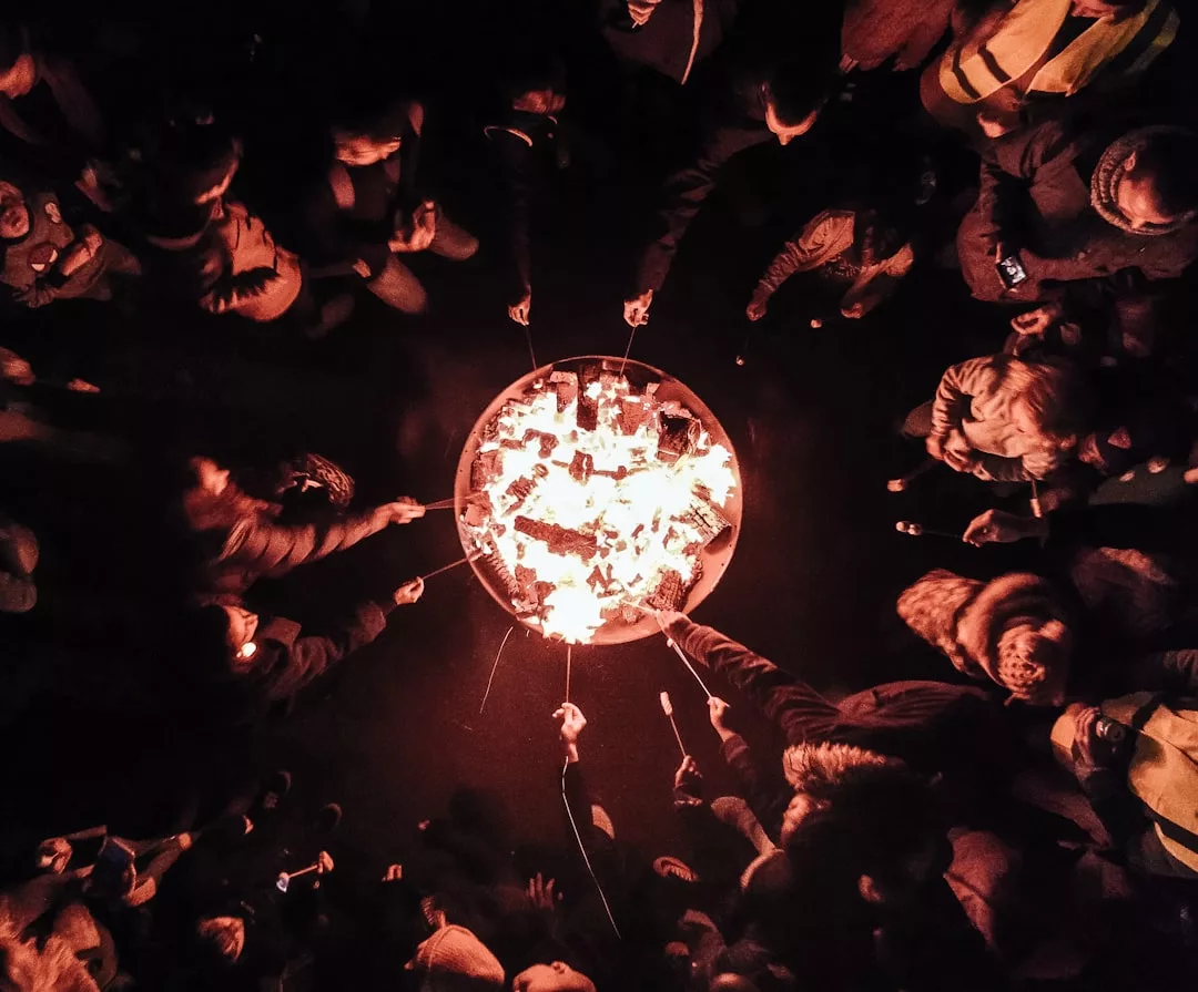 A group of people gather in a circle around a fire pit at night, roasting food on sticks over the flames, enjoying the cozy Glamesia lifestyle.