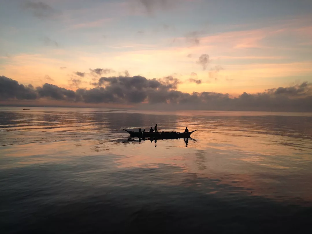 A small boat with people drifts on calm water at sunset, where clouds and soft colors reflect a sense of luxury across the tranquil surface.