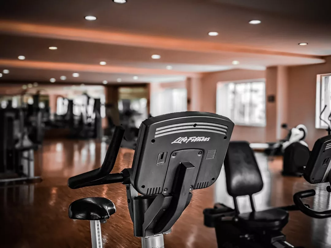 A stationary exercise bike in the foreground of a modern gym with wooden floors and large windows, showcasing a lifestyle of health and luxury; other fitness equipment is visible in the background.