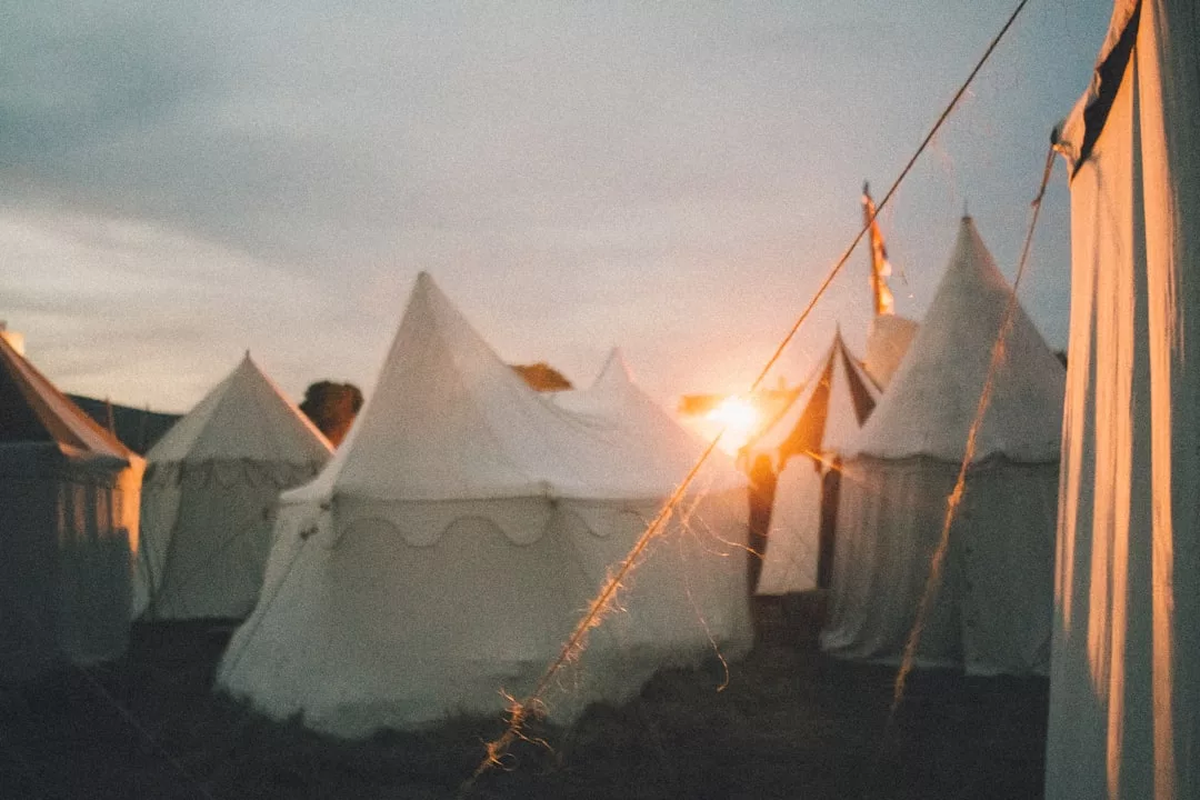 A group of white canvas tents from Glamesia is set up outdoors at sunset, with sunlight casting a warm glow and the luxury tents arranged closely together.