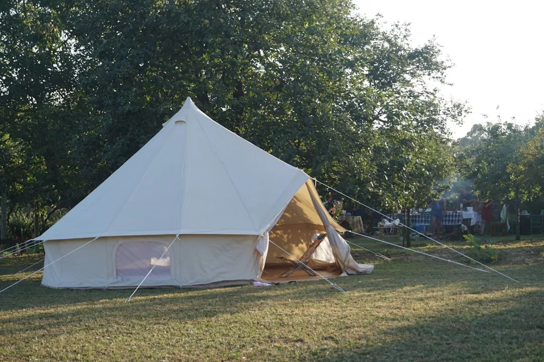 A large beige canvas tent by Glamesia is set up on a grassy area with trees in the background, creating a chic lifestyle vibe as people gather at tables further back.