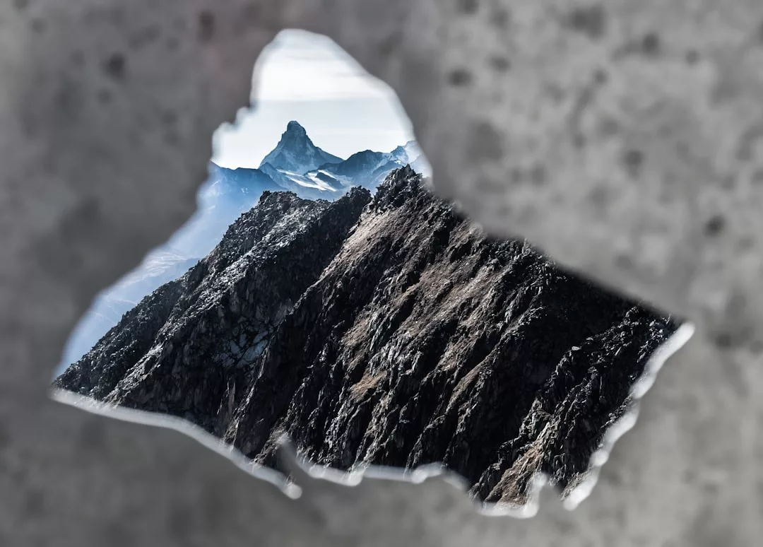 Jagged mountain peaks are visible through a roughly triangular hole in a gray concrete wall, framing the distant summit with a touch of rugged luxury.