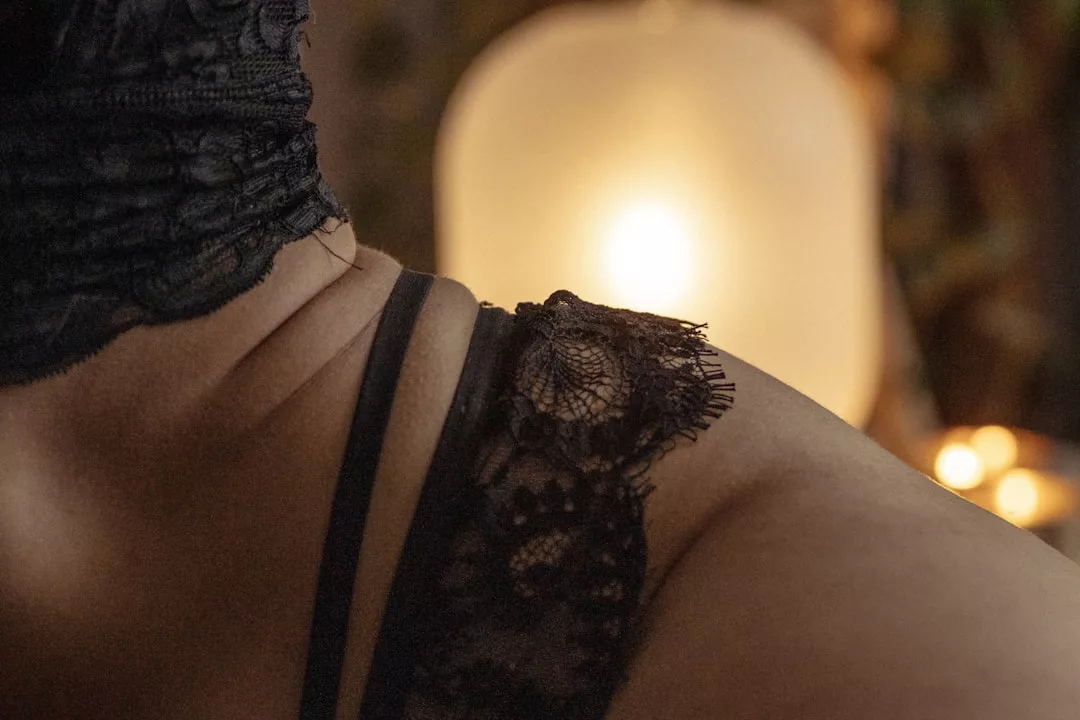 A close-up of a person's shoulder wearing black lace lingerie, with a soft light and blurred lamp in the background, capturing a touch of Laxary lifestyle.