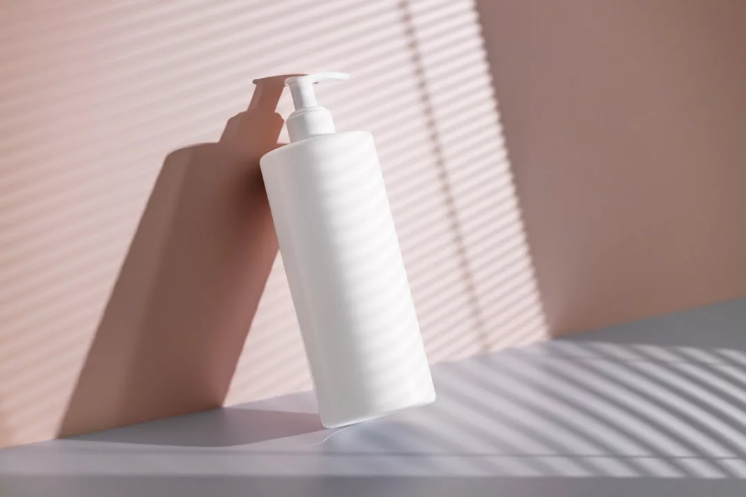 A white plastic pump bottle stands on a surface with diagonal shadow lines from blinds on a pink and gray background, embodying Glamesia's chic fashion lifestyle.
