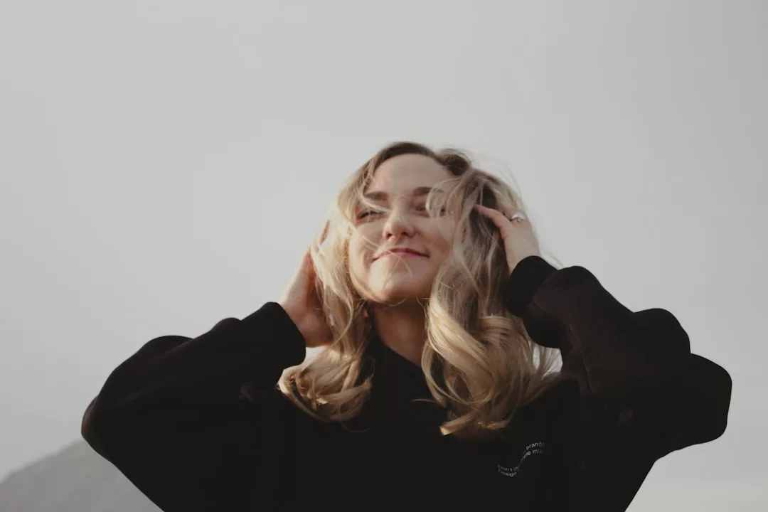 A woman with blonde hair wearing a black sweater stands outdoors, touching her hair and smiling slightly, exuding effortless fashion beneath a cloudy sky in the background.