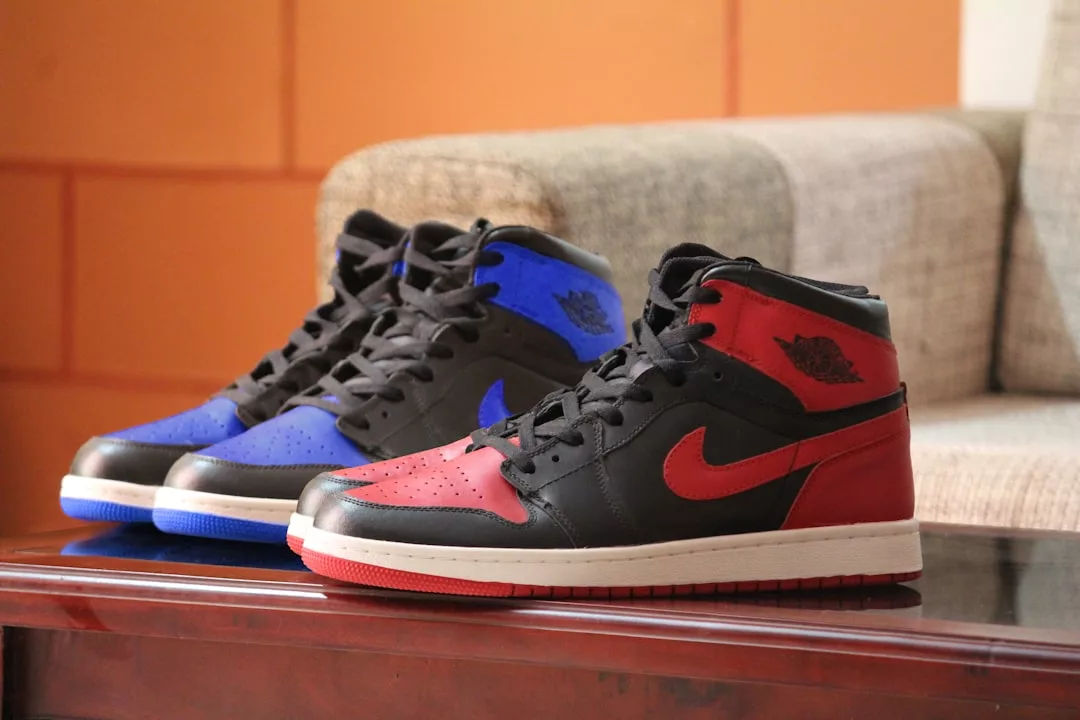 Two pairs of Nike Air Jordan 1 sneakers, perfect for any fashion-forward lifestyle, are displayed on a wooden table; one pair is black and blue, the other is black and red. Discover iconic Glamesia style with every step.