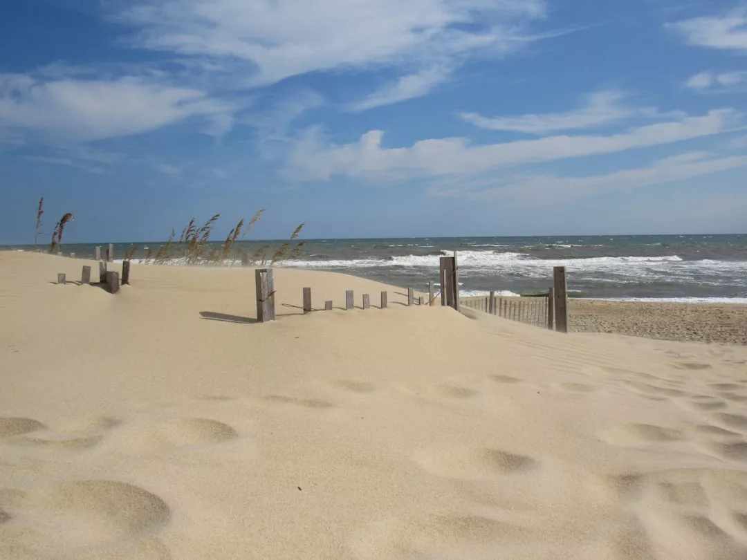 Experience the Glamesia lifestyle on a sandy beach with a wooden fence, sea oats, and ocean waves under a partly cloudy blue sky.