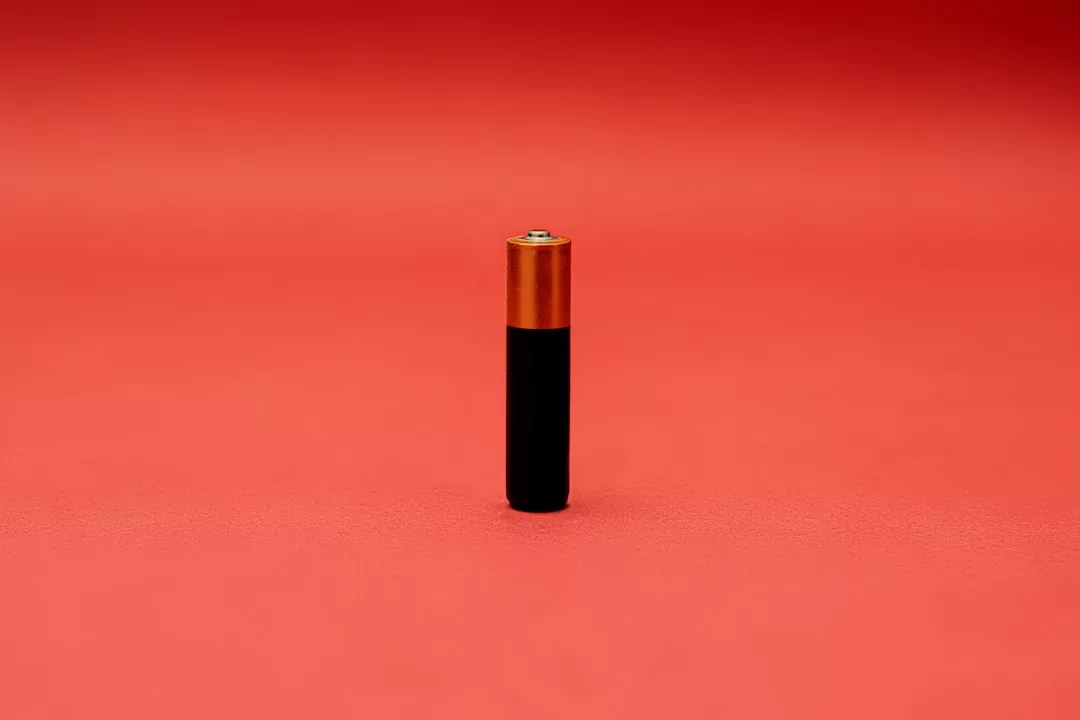 A single AA battery with a copper top and black body stands upright on a flat, textured red surface against a red background, evoking a sense of luxury and modern fashion.