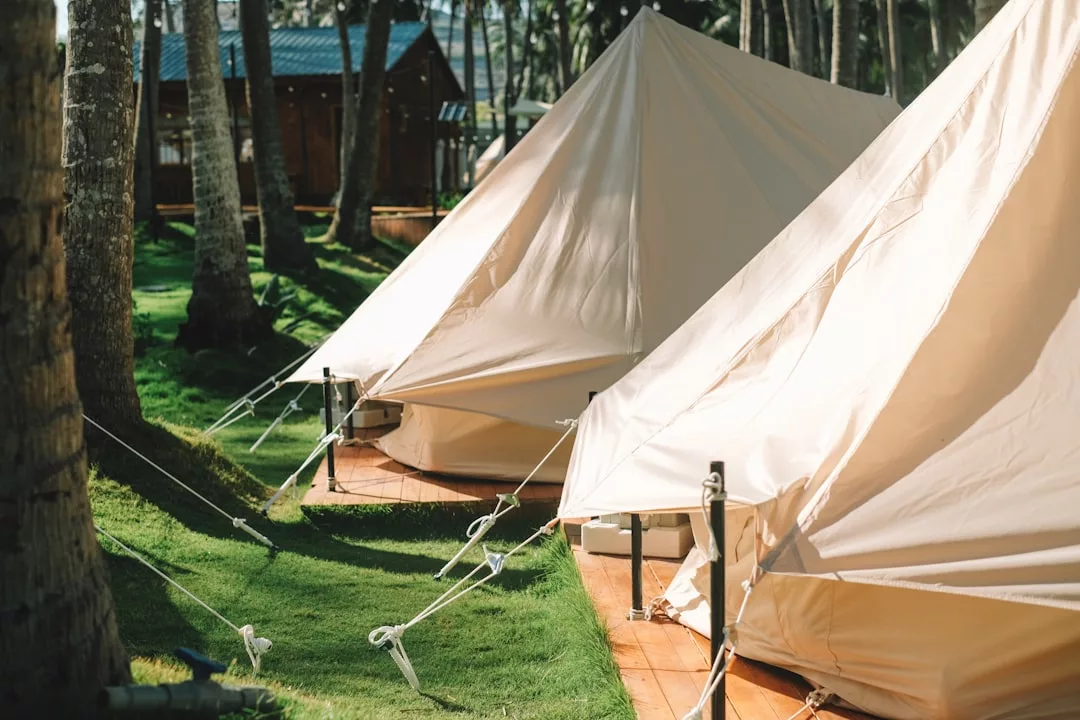 Two beige canvas tents are set up on wooden platforms in a grassy, tree-lined area, offering a taste of the Glamesia lifestyle, with a wooden building visible in the background.