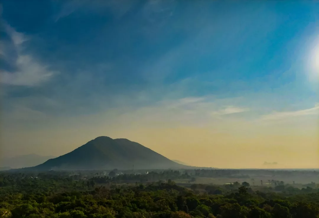 A solitary mountain rises above a green forest under a clear blue sky, with the sun shining brightly on the right side—a perfect glimpse of Glamesia's luxury lifestyle in harmony with nature.