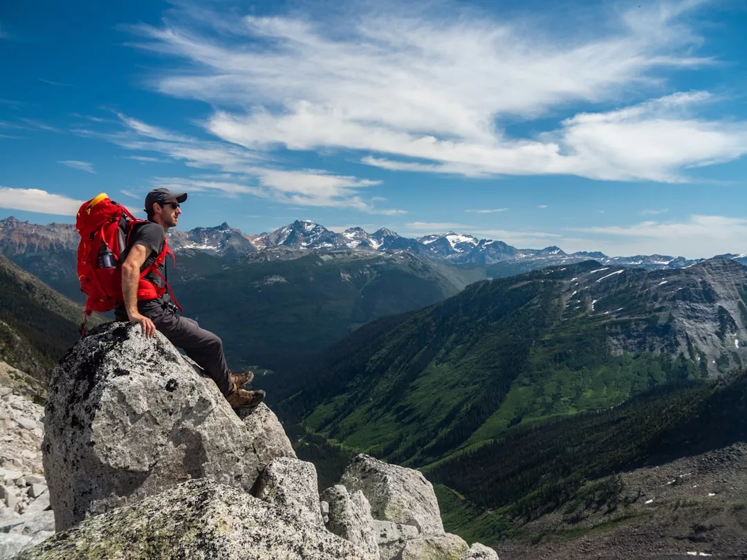 A person wearing a fashionable backpack sits on a large rock, overlooking a scenic mountain landscape under a partly cloudy sky.