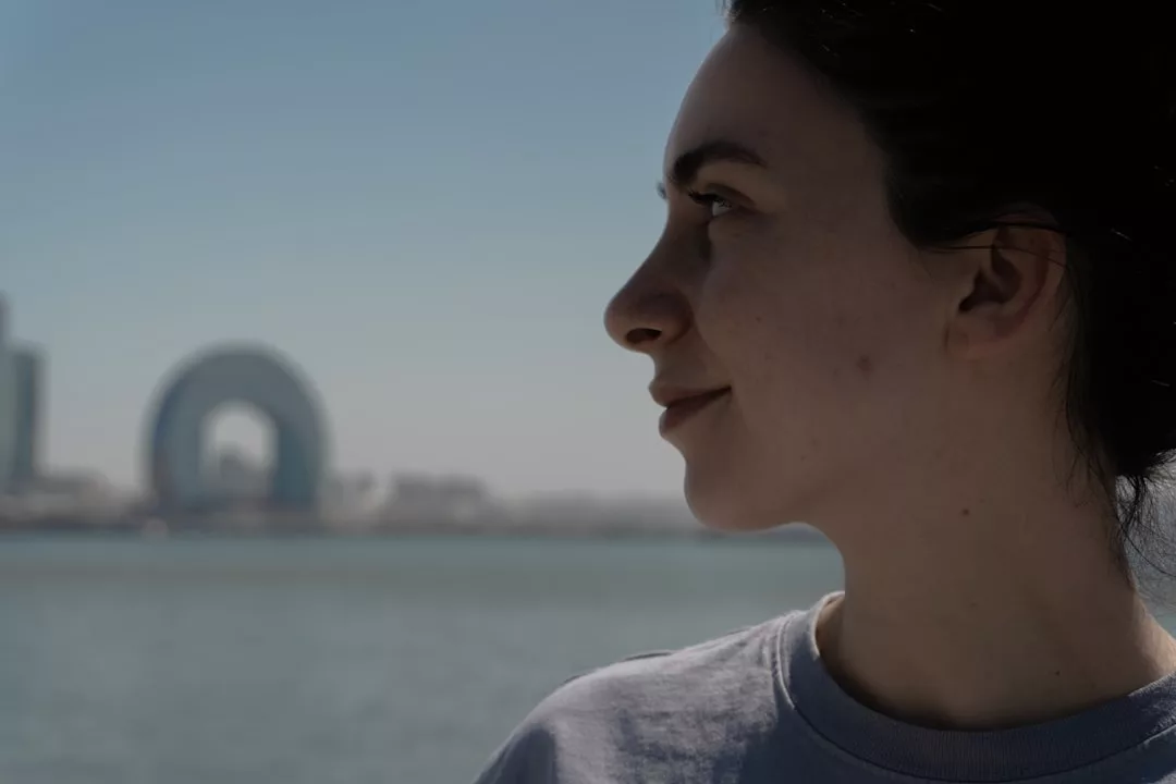 A person in profile looks out over a body of water, with a cityscape and Glamesia's iconic circular building in the background under a clear sky, capturing a moment of modern lifestyle.