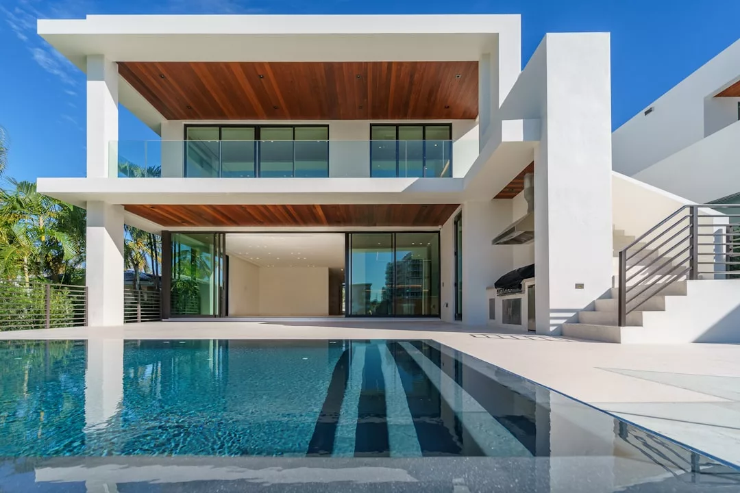 Modern two-story house with large glass windows, wood-paneled ceilings, a balcony, outdoor grill area, and a swimming pool in the foreground under a clear blue sky—perfect for enjoying the Glamesia lifestyle of luxury.