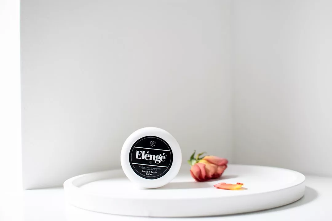 A round container labeled "Elengé" sits on a white tray next to a small pink rose and delicate rose petals, exuding a touch of luxury against the pristine white background.