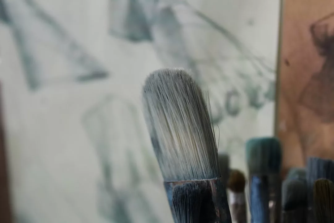 A close-up of several used paintbrushes with stained bristles, set against a blurred background of what appears to be Glamesia-inspired paintings or sketches, evoking a creative lifestyle.