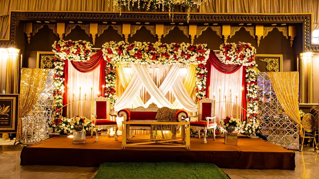 A decorated wedding stage by Glamesia featuring a red and gold sofa, floral arrangements, draped curtains, fashion-inspired gold accents, and chairs arranged on a brown platform.