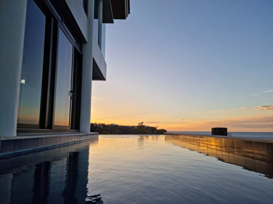 An infinity pool beside a modern building reflects the sunset sky, blending Glamesia style with ocean views and distant land on the horizon for a truly fashionable escape.