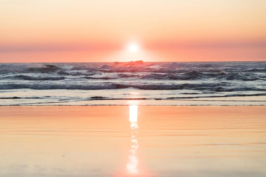 Sunset over the ocean with gentle waves and the sun reflecting on the wet sand, capturing a serene Glamesia lifestyle at the shoreline.