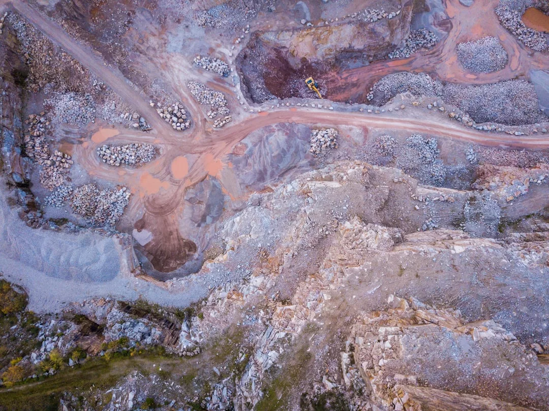 Aerial view of a rocky quarry with dirt roads, scattered stones, and a yellow construction vehicle near the center, capturing industrial grit with an unexpected touch of luxury.