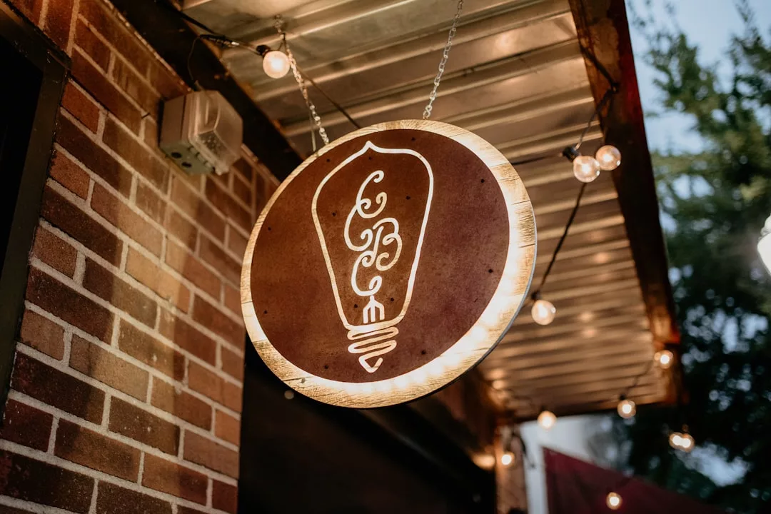 A round wooden sign with a white lightbulb design hangs outside a brick building under a metal awning with string lights, adding a touch of Glamesia fashion to the scene.