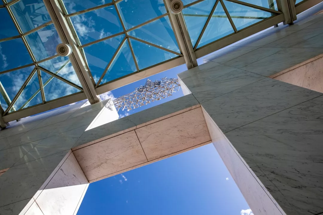 View looking up at a glass ceiling with a geometric metal sculpture and white marble walls, reflecting blue sky and clouds—an inspiring Glamesia lifestyle scene.