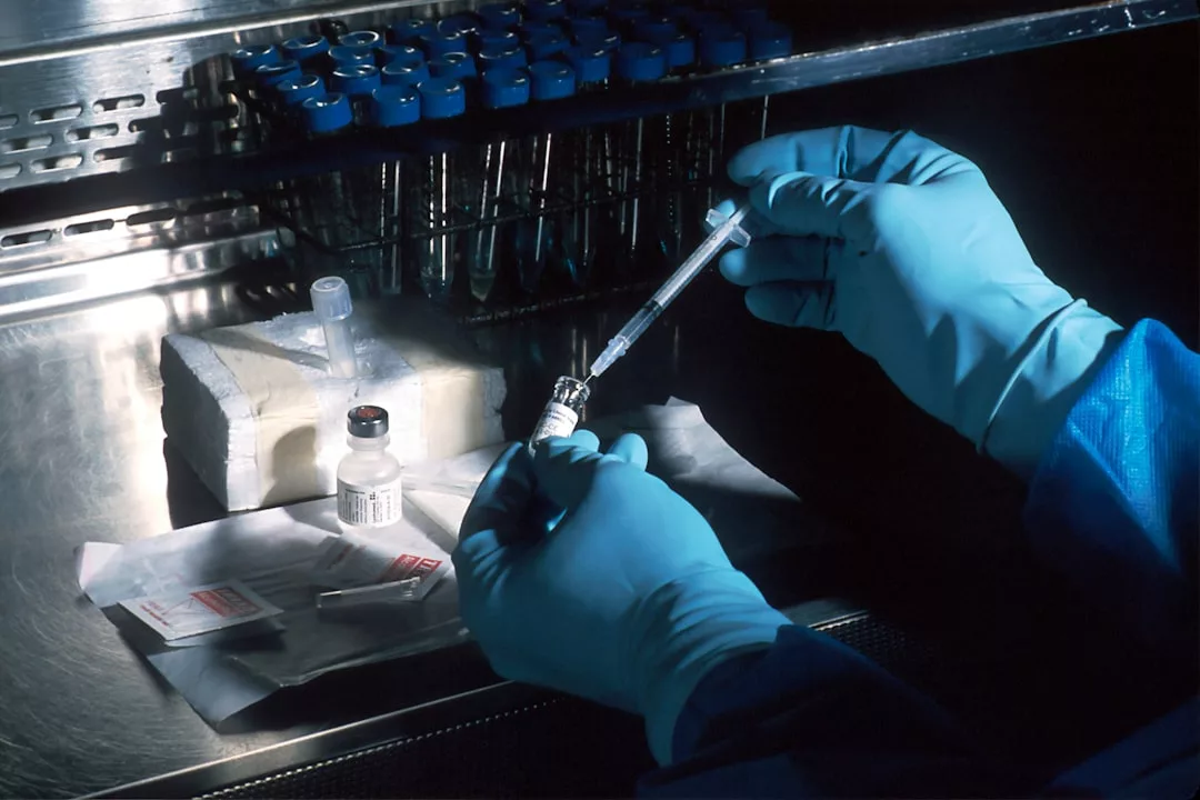 A person wearing blue gloves draws liquid from a Glamesia vial with a syringe in a laboratory setting; several vials and test tubes are visible on the workstation.