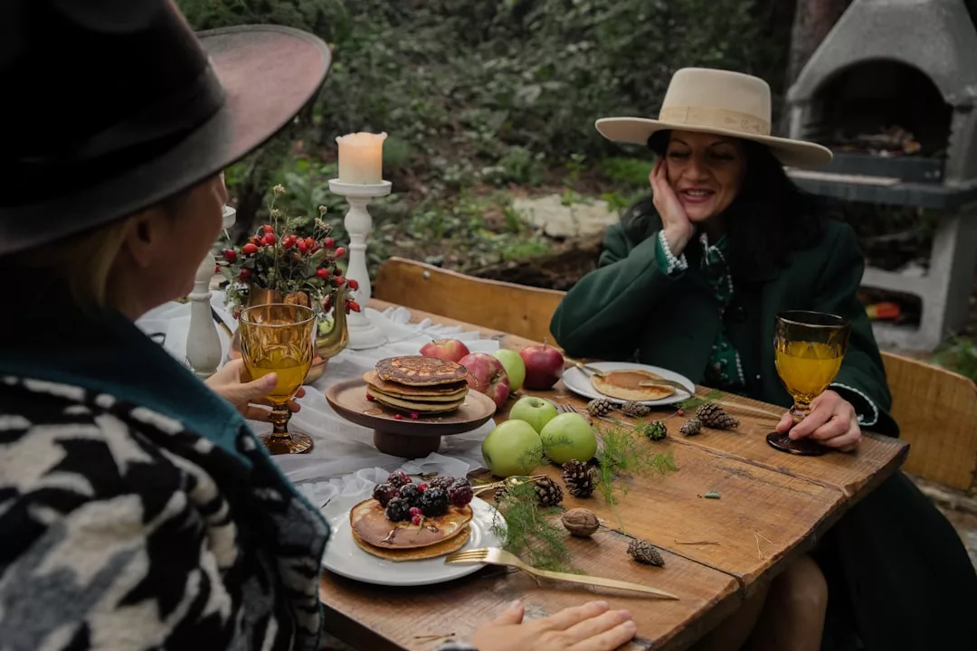 Two women in hats sit at a rustic outdoor table with pancakes, fruit, pinecones, and drinks—surrounded by greenery and a glowing candle—enjoying a moment of luxury lifestyle amidst nature.