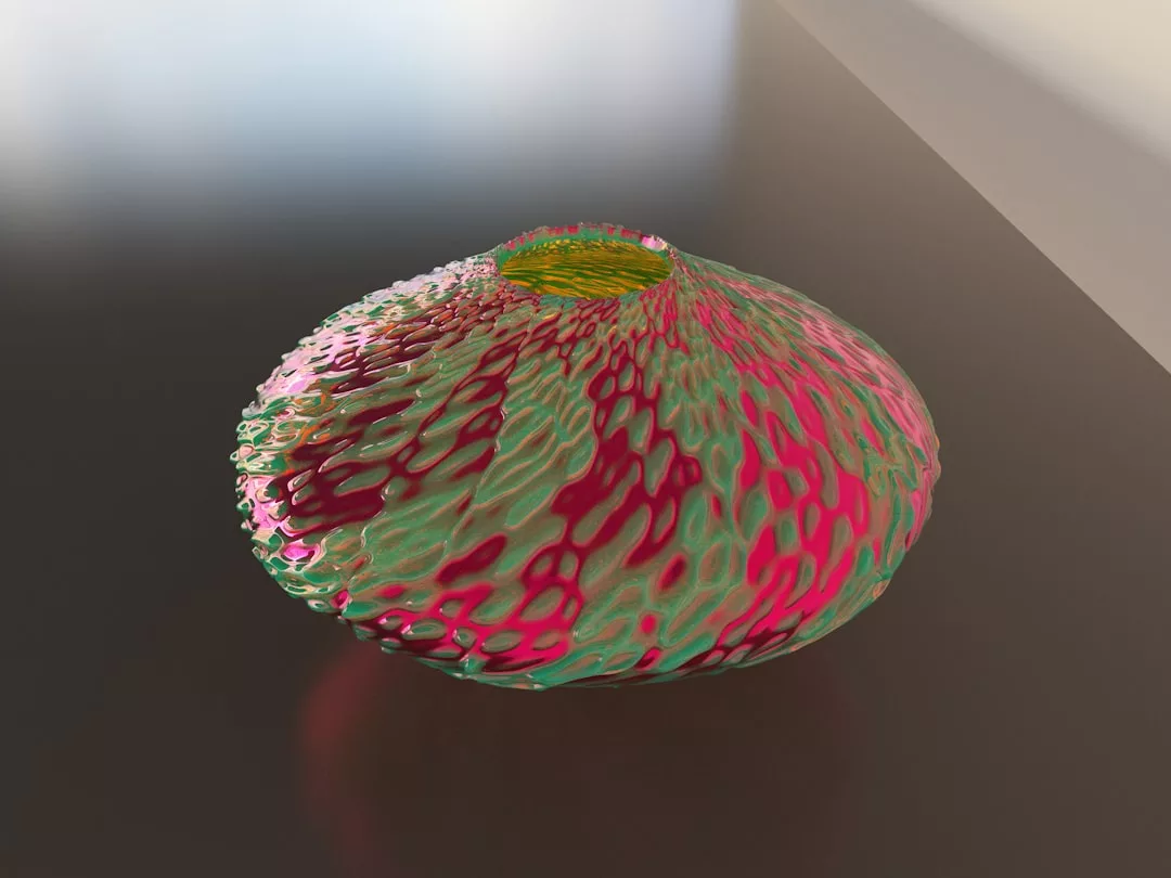 A textured, round Glamesia vase with a narrow opening, covered in green, pink, and red patterns, sits on a reflective, dark surface with soft lighting.