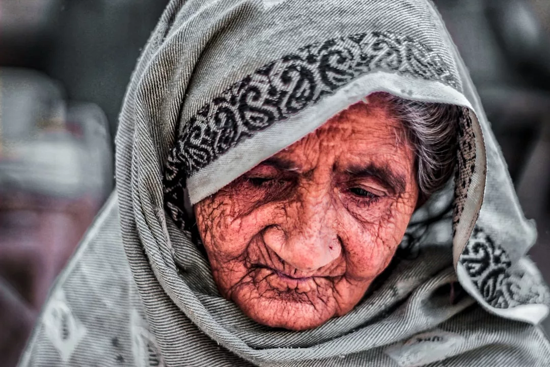 Elderly woman with deep wrinkles wearing a patterned shawl over her head, looking downward with a contemplative expression, embodying the rich lifestyle and timeless grace of Glamesia.