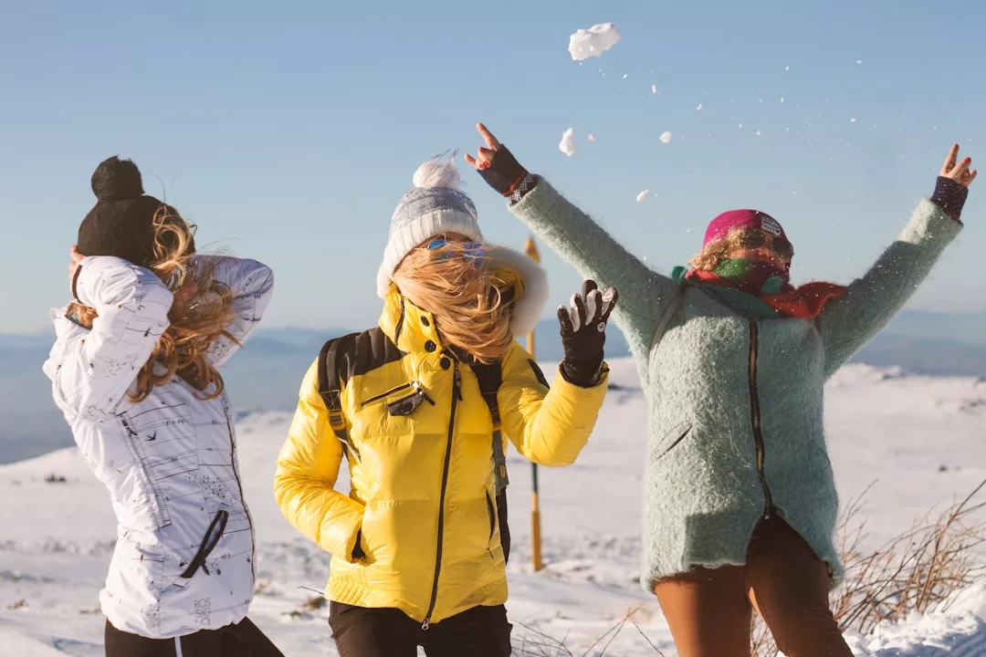 Three people in winter jackets showcase their fashion and lifestyle as they stand outside in a snowy landscape; one throws snow into the air while the others react to the wind.