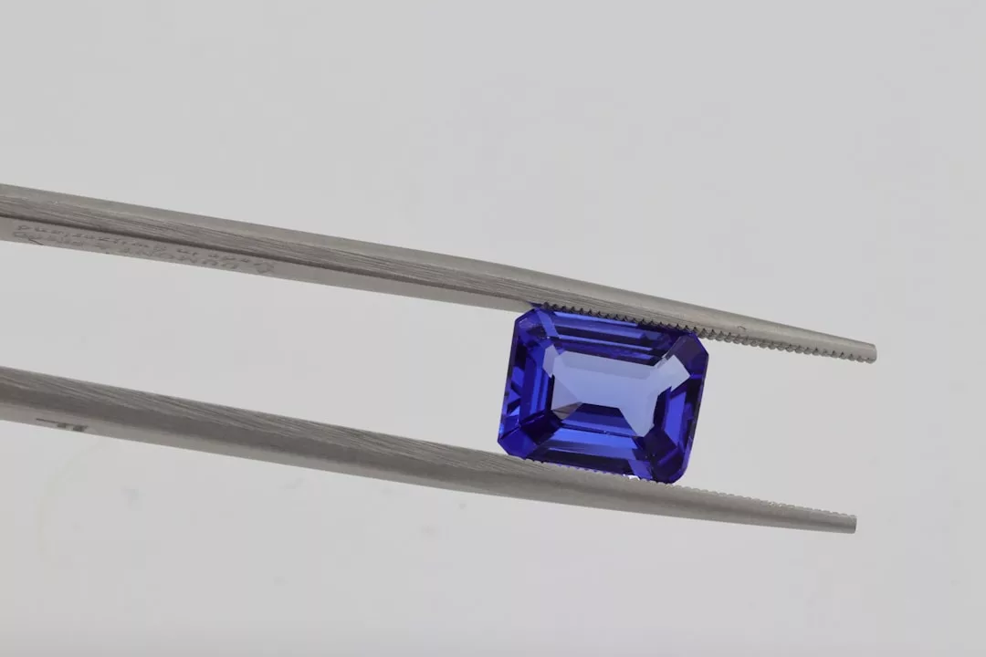 A pair of tweezers holding a rectangular, emerald-cut blue gemstone against a plain, light grey background, capturing the essence of luxury and refined lifestyle.