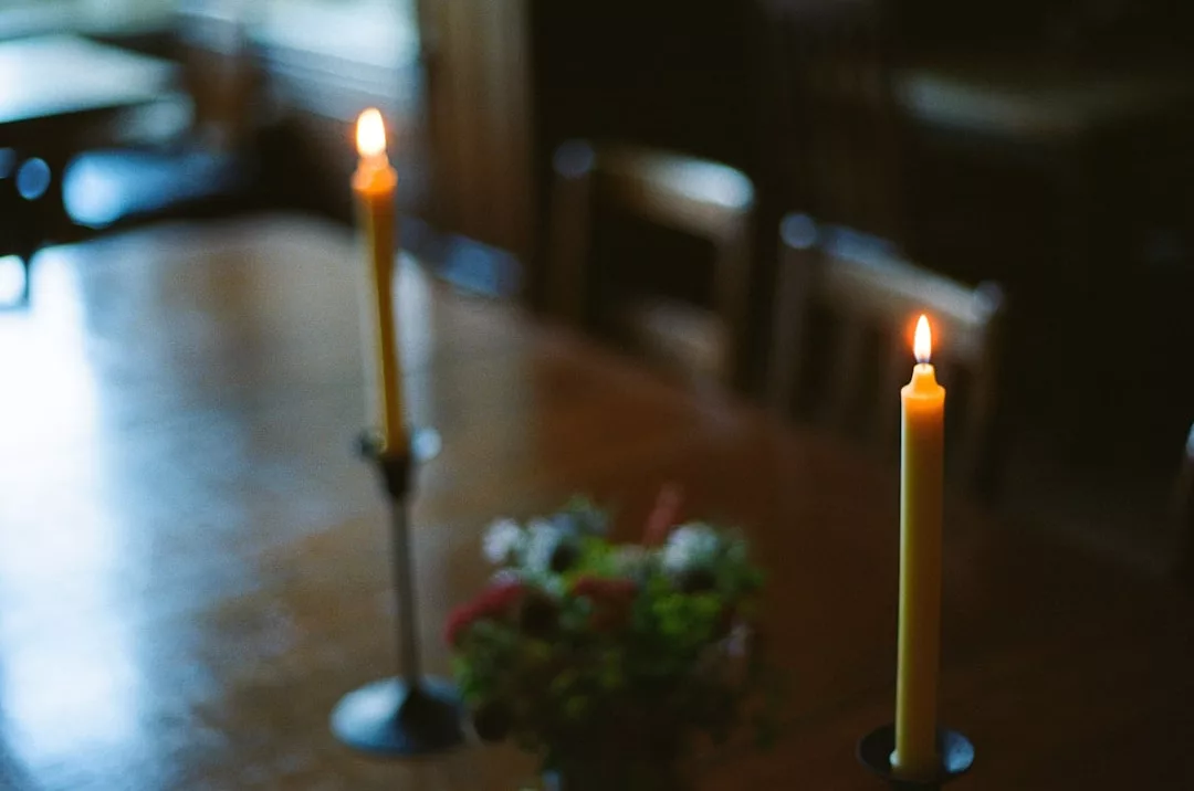 Two lit candles in holders on a wooden table with a small floral centerpiece create a sense of luxury; chairs and a window are visible in the blurred background, evoking an elegant lifestyle setting.