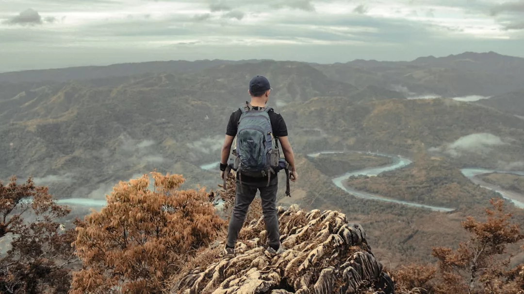 A person wearing a backpack stands on a rocky outcrop, overlooking a large valley with winding rivers and distant hills under a cloudy sky, capturing the untamed beauty of Glamesia.