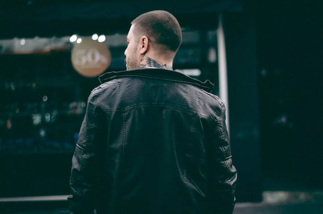 A man with short hair and a neck tattoo wears a black leather jacket, embodying urban fashion as he stands with his back to the camera in a city setting.