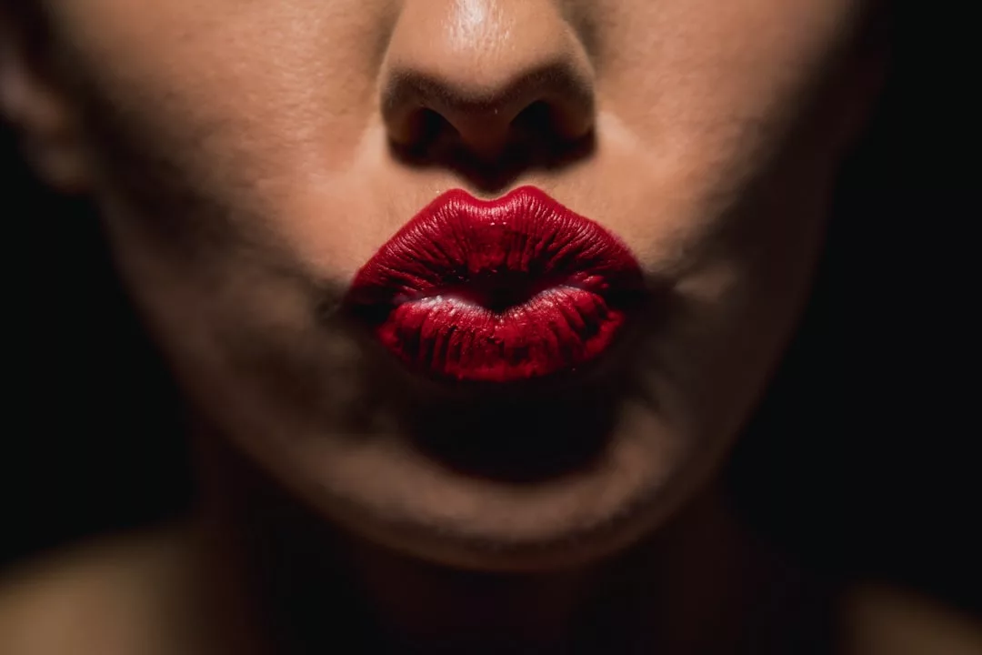 Close-up of a person with red lipstick pursing their lips in a kiss shape against a dark, luxury-inspired background.