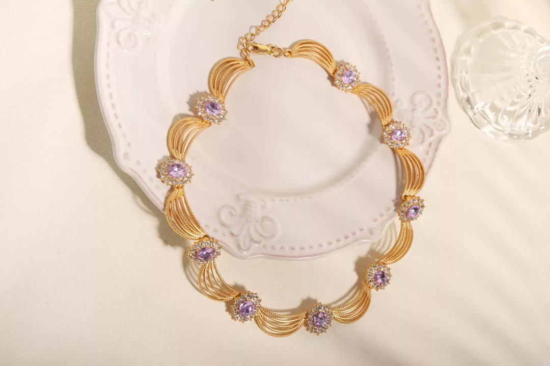 A gold necklace with purple gemstone accents exudes luxury as it rests on a white decorative plate, with a glass object nearby.