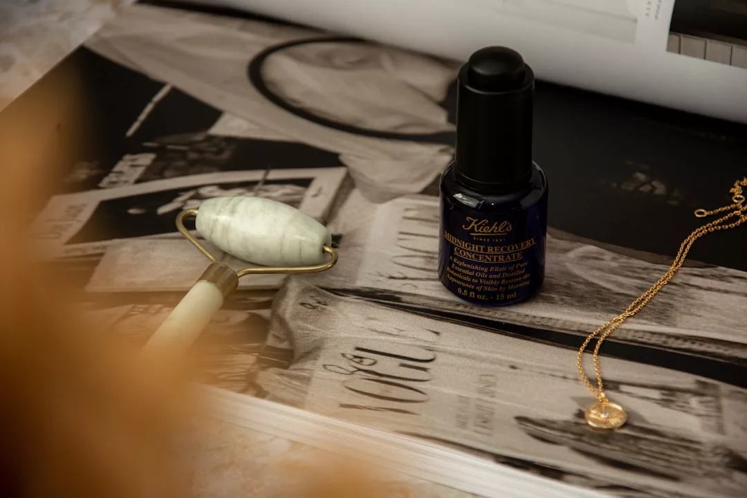 A jade facial roller, a bottle of Kiehl's Midnight Recovery Concentrate, and a gold necklace from Glamesia rest on an open magazine with black-and-white images, evoking a sense of effortless luxury.